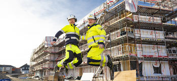 To menn i arbeidsklær hopper foran bygg under oppføring.